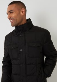 Schwarze gepolsterte Jacke mit hohem Kragen, ausgestattet mit zwei Fronttaschen, Druckknöpfen und einer gesteppten Textur. Widerstandsfähiges und warmes Design.