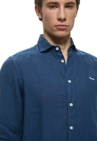Camicia di lino blu con colletto button-down, cinque bottoni e un piccolo logo di un cane bianco sul lato sinistro del petto. Tessuto liscio e strutturato.