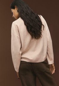 Kvinde med langt mørkt hår iført en lys pink sweater og mørke bukser, stående sidelæns mod en brun baggrund.