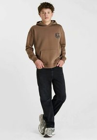 Bruine capuchontrui met een voorkantzak en bedrukt logo, gecombineerd met donkere spijkerbroek en grijze sportschoenen. Eenvoudige, ontspannen pasvorm.