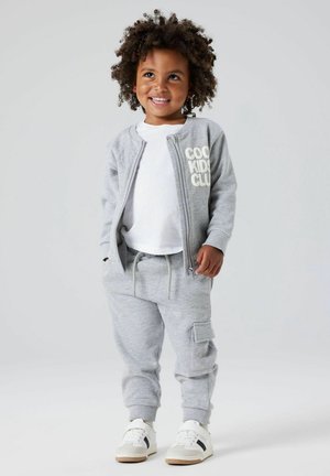 Survêtement en polaire grise avec veste zippée et pantalon cargo, arborant le texte "COOL KIDS CLUB" et un t-shirt blanc, associé à des baskets blanches.