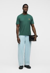 Άνδρας με πράσινο μπλουζάκι με μικρό λεκέ αλεπούς, ανοιχτό μπλε τζιν, μαύρα παπούτσια, κρατώντας μαύρη διπλωμένη τσάντα σε απλό φόντο.