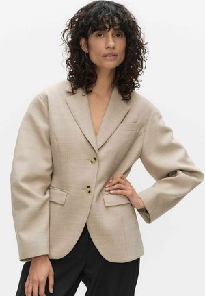 Nainen, jolla on kiharat hiukset, pukeutunut beigeen ylisuurikokoiseen kaksinappiseen bleiseriin ja mustiin housuihin, poseeraa yhdellä kädellä lanteillaan.