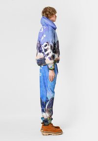 Chaqueta acolchada con gráfico de montaña en colores pastel y texto blanco grande, combinada con pantalones jogger azules con estampado y botas marrones.