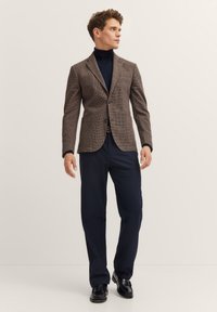 Brauner, gemusterter Blazer mit Reverskragen, getragen über einem marineblauen Rollkragenpullover, kombiniert mit dunklen Hosen und schwarzen Schuhen. Glatte Textur insgesamt.