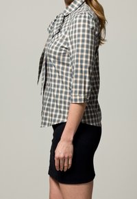 Donna in piedi di lato che indossa una camicia a quadri blu e beige con maniche a tre quarti e una gonna nera aderente.