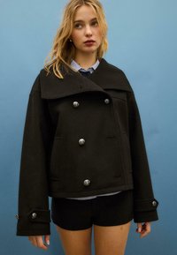 Giacca nera in misto lana con colletto oversize, bottoni doppi in argento e maniche larghe. Indossata sopra una camicia a righe blu e pantaloncini neri.