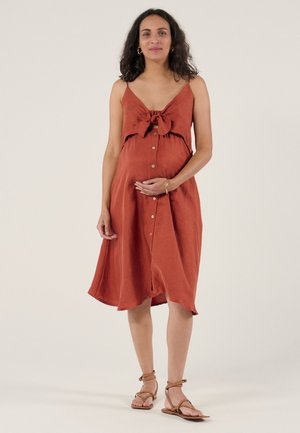 Schwangere Frau in einem ärmellosen, rostfarbenen Kleid mit Knopfdetail und Schleife, barfuß stehend vor hellem Hintergrund.