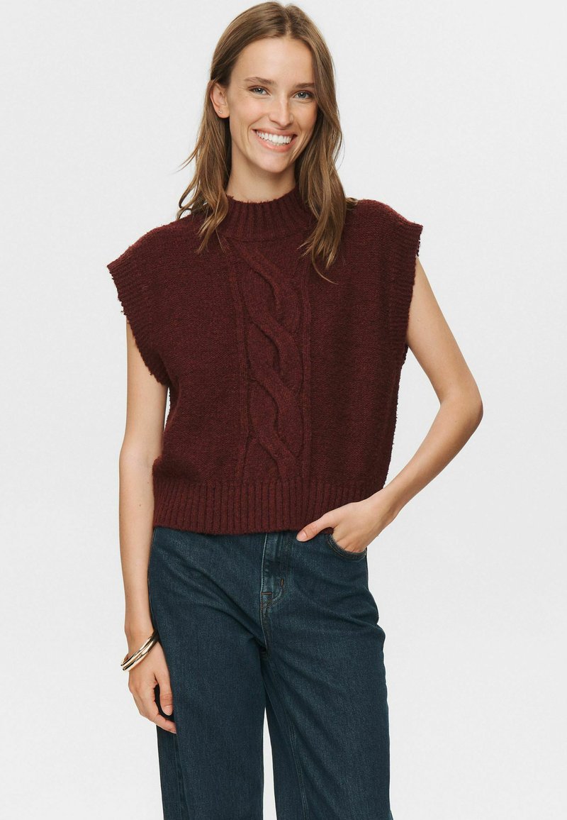 Šypsena moteris, vilkinti be rankovių tamsiai raudoną megztinį su pintinėmis ir tamsiai mėlynomis džinsomis, viena ranka kišenėje ir su apyrankėmis ant riešo.