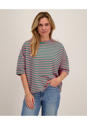 Vrouw met lang blond haar, dragend een groen en roze gestreept oversized T-shirt en blauwe spijkerbroek, één hand in de zak, neutrale achtergrond.