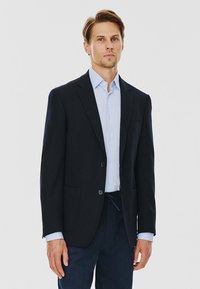 Granatowa marynarka z dwoma guzikami, klapami w szpic i przednimi kieszeniami. Noszona na jasnoniebieskiej koszuli. Gładki materiał z dopasowanym krojem.