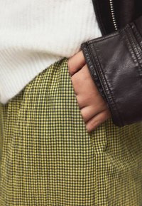 Tissu à carreaux jaune et noir avec une texture lisse ; main dans la poche ; superposé avec un pull blanc côtelé et une veste en cuir noir.