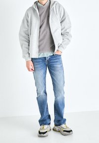 Sudadera con cremallera de color gris claro sobre un sudadera de cuello redondo gris, combinada con jeans de mezclilla azul y zapatillas grises con acentos amarillos.
