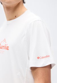 Άτομο που φοράει άσπρο υφασμάτινο πουκάμισο με κοντά μανίκια και κοραλλί λογότυπα Columbia στο στήθος και το μανίκι, μπροστά σε απλό φόντο.