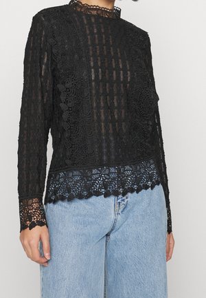 Schwarzes transparentes Spitzenoberteil mit langen Ärmeln, floralen Mustern und gewellten Kanten, kombiniert mit hellblauen Jeans.