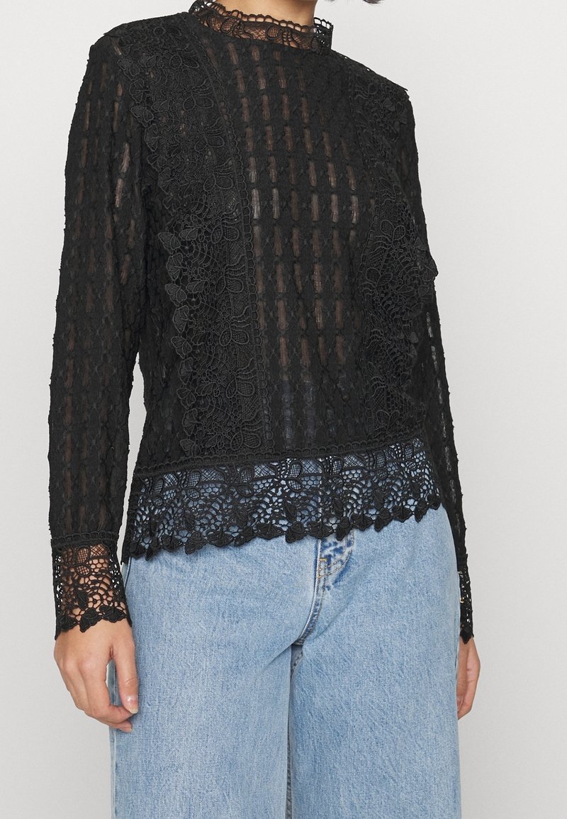 Haut noir en dentelle transparente à manches longues avec motifs floraux et bords festonnés, porté avec un jean en denim bleu clair.