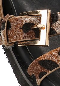 Sandalia de tira con estampado de animal en tonos marrones y hebilla de metal dorada sobre una suela negra texturizada. La tira presenta una textura suave similar al pelo.