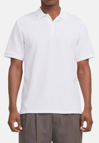 Polo shirt blanc en tissu texturé, avec un col, des manches courtes et un logo discret sur la poitrine gauche.