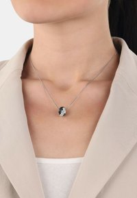 Silverhalsband med en svart och vit sammanflätad berlock, med släta och texturerade ytor, bärs över en ljus tanktopp.