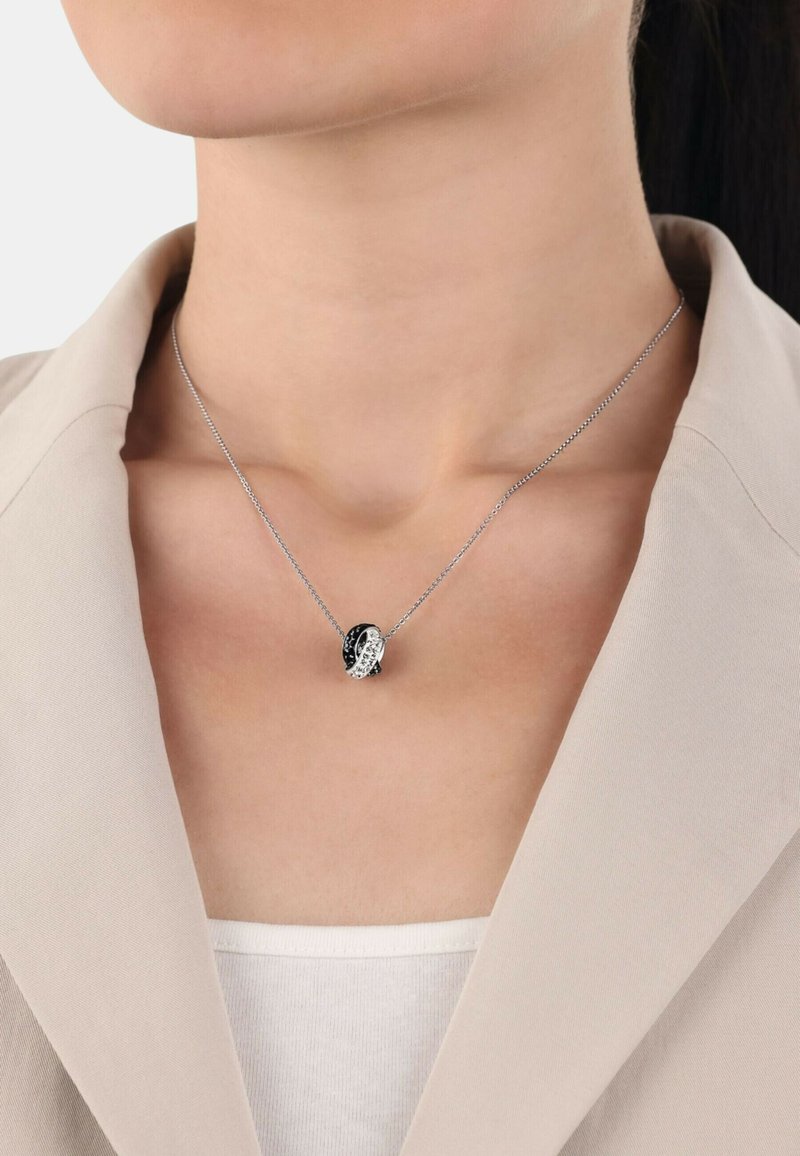 Silverhalsband med en svart och vit sammanflätad berlock, med släta och texturerade ytor, bärs över en ljus tanktopp.