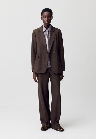 Blazer lungo marrone con due bottoni, abbinato a pantaloni larghi coordinati. Sotto, una camicia a righe e dettagli di pantofole decorate.