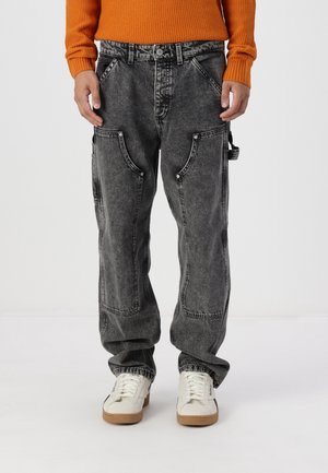 Mężczyzna ubrany w czarne spodnie cargo w stylu acid wash z wieloma kieszeniami, białe trampki oraz pomarańczowy teksturowany sweter, stojący na jednolitym tle.