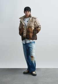 Καφέ και μπεζ καρό μπουφάν με φερμουάρ μπροστά και υπερμεγέθη εφαρμογή, σε συνδυασμό με ανοιχτό μπλε τζιν με φθορές και λεπτομέρειες με κολάζ.
