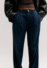 Personne portant un jean plissé taille haute bleu foncé, un top court marron et une veste en cuir noire, les mains dans les poches.