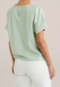 Ljusgrön kortärmad blus med en nyckelhålsdetalj i ryggen, tillverkad av mjukt tyg. Bärs med vita jeans med bakfickor.