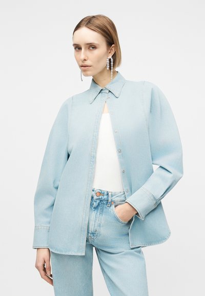 Γυναίκα που φοράει ανοιχτό μπλε φαρδιά τζιν μπουφάν πάνω από λευκό τοπ, σετ με ανοιχτό μπλε τζιν παντελόνι και μακριά κρεμαστά σκουλαρίκια, ποζάρει με το χέρι στην τσέπη.