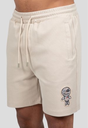 Beige cotton shorts with an elastic waistband, featuring a front pocket and a multicolored cartoon character embroidery on the left leg.