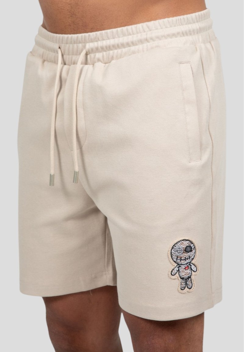 Bermudas de algodón beige con cinturilla elástica, que cuentan con un bolsillo delantero y un bordado de un personaje de dibujos animados multicolor en la pierna izquierda.