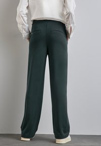 Celana lebar hijau tua dengan tekstur halus, dilengkapi dengan saku belakang dan waistband tinggi, dipadukan dengan kemeja putih berlengan panjang.