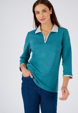 Camisa de manga larga en color azul verdoso con escote en V, que presenta un cuello y puños con estampado floral. Incluye un bolsillo en el pecho y un corte holgado.