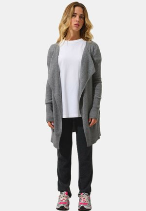 Cardigan grigio chiaro con apertura davanti, maniche a coste, design a strati; indossato sopra una maglietta bianca, abbinato a pantaloni scuri e sneakers.