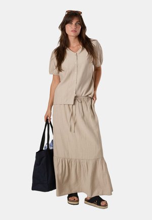 Woman in beige button-up short-sleeve top and matching long skirt holds black tote bag, wearing black platform sandals and sunglasses on head.