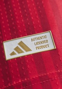 Χρυσό λογότυπο Adidas και κείμενο "Authentic Licensed Product" σε λευκή ετικέτα που είναι προσαρτημένη σε υφασμάτινο υλικό με υφή κόκκινου χρώματος.