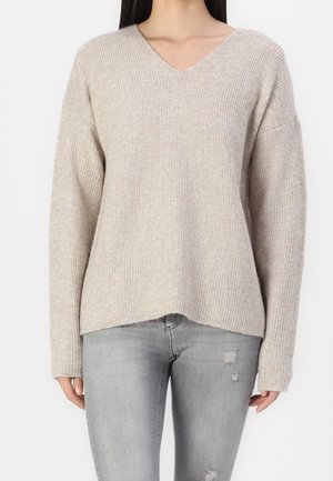 Femme portant un pull beige côtelé à col en V et un jean skinny gris clair déchiré, debout devant un fond blanc uni.