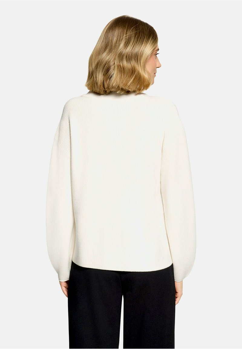 Femme aux cheveux blonds mi-longs, portant un pull blanc à manches longues et un pantalon noir, dos tourné sur un fond uni.