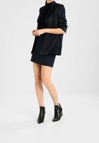 Blazer de satén negro con mangas enrolladas sobre una blusa negra ajustada, combinado con una falda mini de textura azul marino y botines negros de cuña.