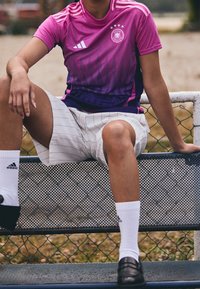 Różowa i fioletowa sportowa koszulka z wzorem marki Adidas w połączeniu z białymi szortami. Czarne buty i białe skarpetki. Siedzi na siatkowej ławce przy ogrodzeniu.