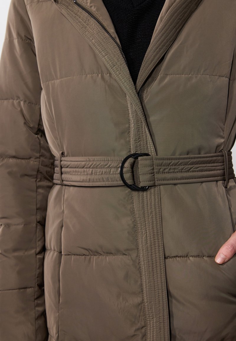 Manteau matelassé vert olive avec fermeture éclair, taille ceinturée avec une boucle ronde noire, et détails de couture verticale sur les côtés.