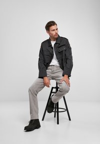 Chaqueta negra, ligera y con múltiples bolsillos, llevada sobre una camisa blanca. Jeans grises y botas negras completan el conjunto. Sentado en un taburete.