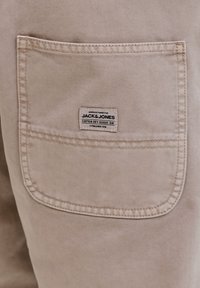 Poche en tissu beige sur un pantalon avec une étiquette rectangulaire indiquant "Fabriqué pour JACK & JONES Cotton Dry Goods Div. Fondée en 1990."