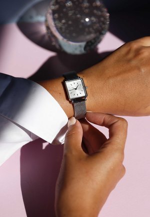 Zilveren rechthoekige horloge met een mesh-band om een pols, met een witte wijzerplaat, zwarte minutenmarkers en een datumschijf, op een roze oppervlak.