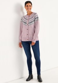 Kvinna som ler, iklädd en rosa mönstrad kofta, mörkblå skinny jeans och svarta ankelskor, stående mot en vit vägg.