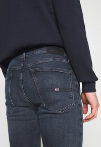 Vaqueros de mezclilla azul oscuro con un ajuste de tiro medio. Presenta bolsillos traseros con un parche de logo y costuras en contraste. Textura de tela suave.