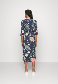 Vestido floral azul-marinho com mangas de três quartos, padrões florais em rosa e verde,laço na cintura e fendas laterais. Usado com sandálias brancas de plataforma.
