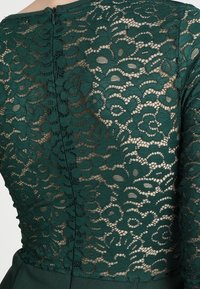 Tissu en dentelle florale vert foncé avec une fermeture éclair au dos, révélant la peau sous le motif complexe de la dentelle.