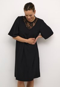 Schwarzes Kleid mit V-Ausschnitt und Spitzendetails, kurzen Ärmeln und gerippter Textur. Der Saum endet oberhalb des Knies.
